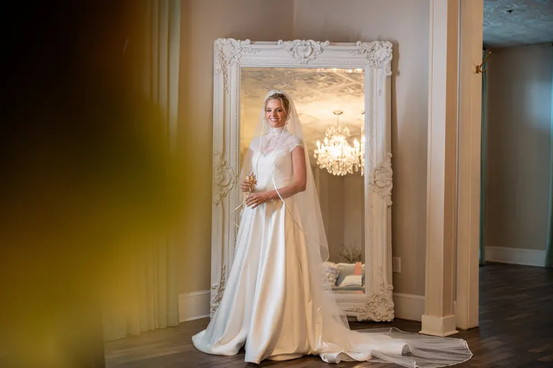 Person in a flowing white gown stands before an ornate mirror in an elegant interior space.