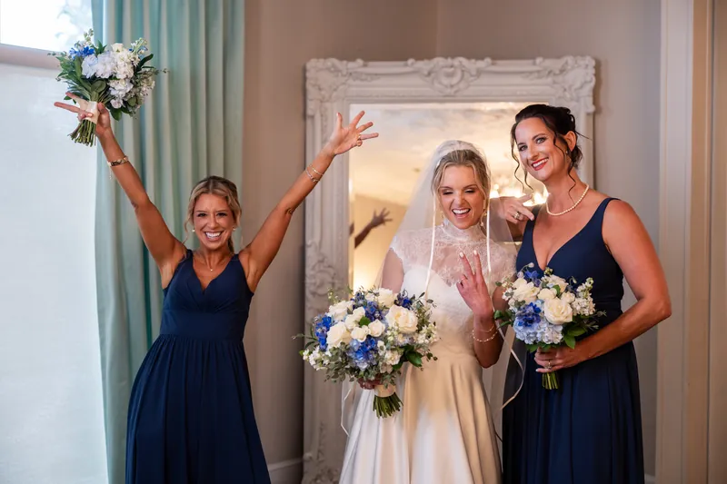 Three people in formal attire hold wedding bouquets, with one on the left raising their arms in celebration.