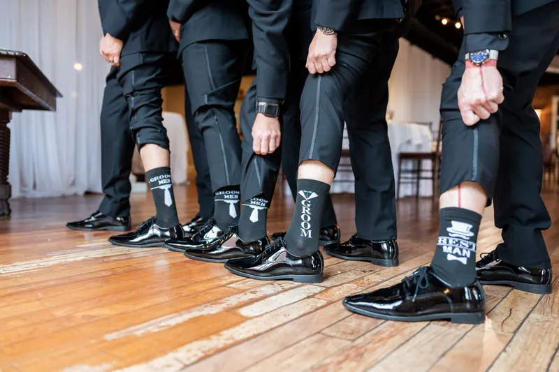 Group of five people in black suits and dress shoes stand together on a wooden floor, displaying patterned socks.