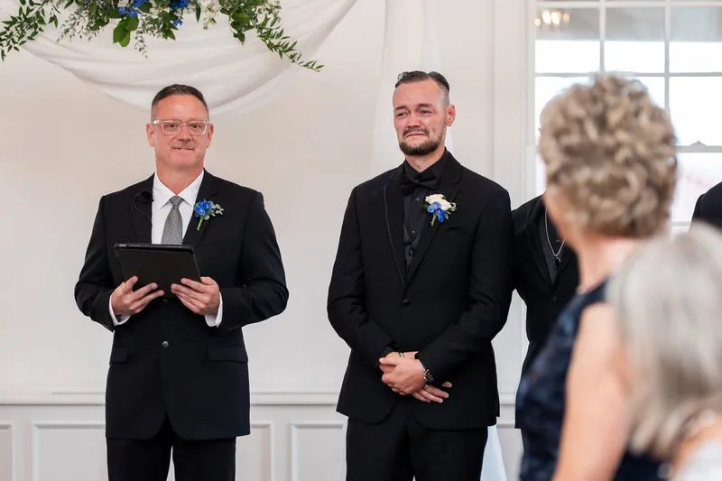 Two people in dark suits with blue boutonnieres stand at an altar facing guests during a ceremony.