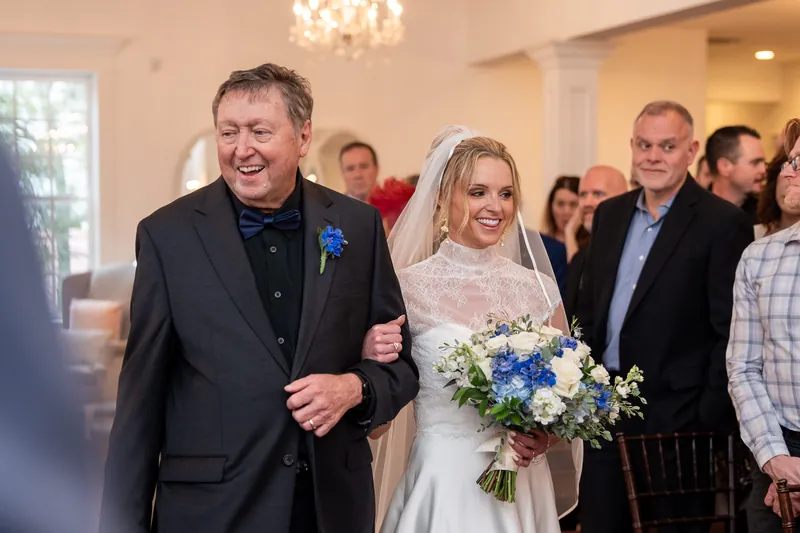 A person in a dark suit walks beside someone in a white wedding gown and veil, holding a blue and white bouquet.