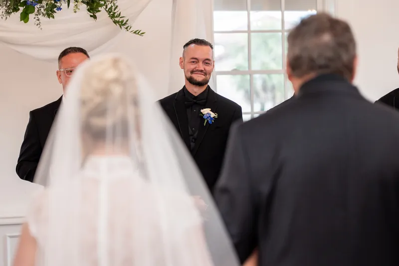 Person in a white gown and veil stands before two people in dark suits during an indoor ceremony.