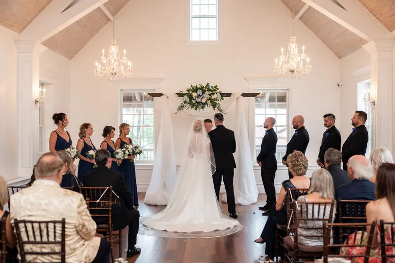 A person in a white gown with long train walks down the aisle as guests seated on both sides watch during a chapel ceremony.