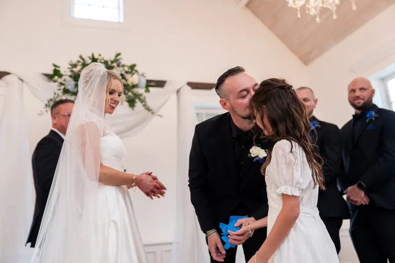 Two people in wedding attire kiss while standing near a floral arch, surrounded by family members in formal wear.