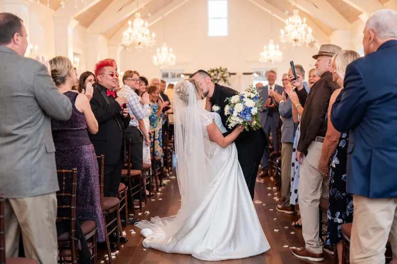 Wedding guests stand and applaud as a couple in white gown and black suit walk together down the center aisle of a brightly lit chapel.