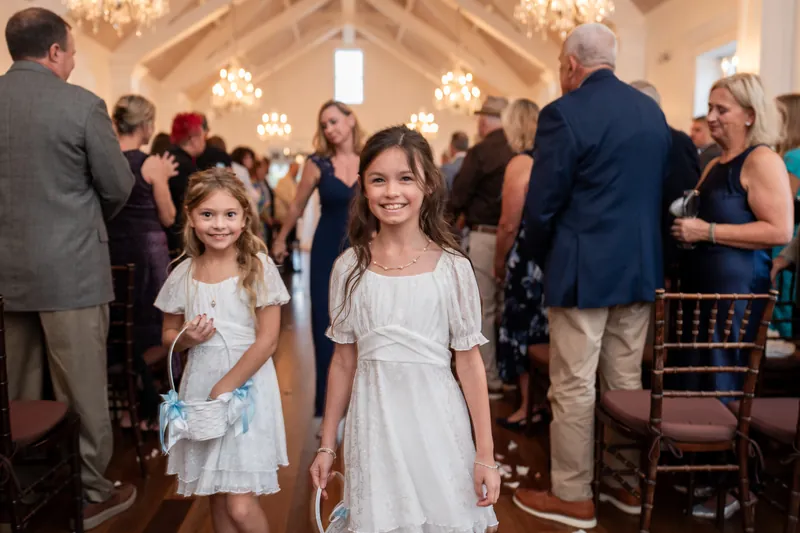 Two young girls in white dresses walk down the aisle of a chapel filled with standing guests in formal clothing.
