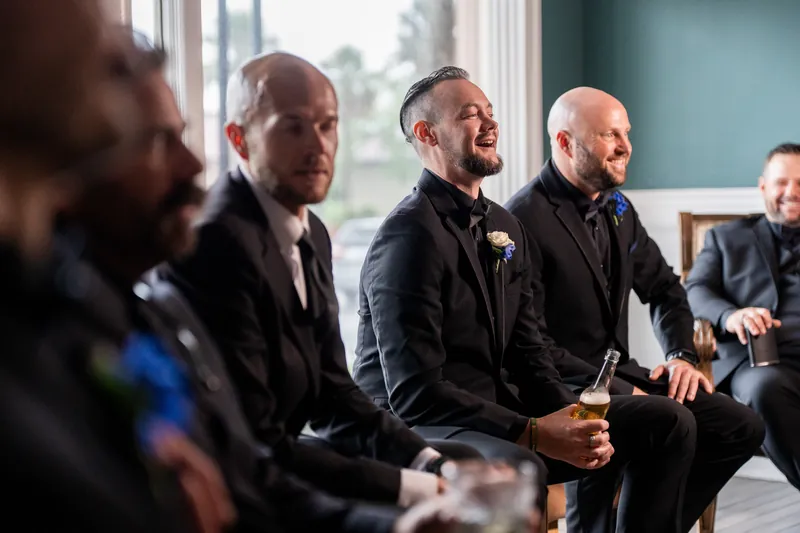 Several men in dark suits stand together indoors near tall windows, one wearing a boutonnière and smiling.