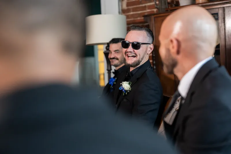A man in sunglasses and a dark suit with a white boutonnière laughs while talking with others in an elegant interior space.