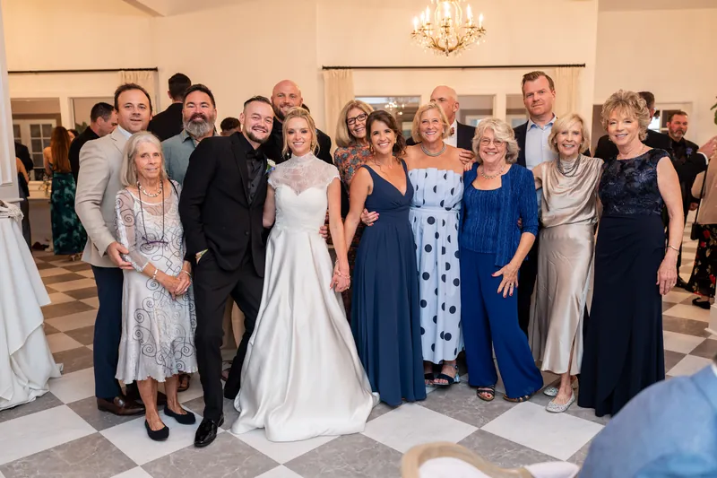 Li and Cody stand centered in a formal group photo with family members in an elegant ballroom with a crystal chandelier and checkered floor.