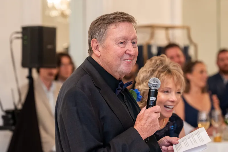 A man and woman hold a microphone while speaking at a reception, surrounded by seated guests in a cream-colored venue.