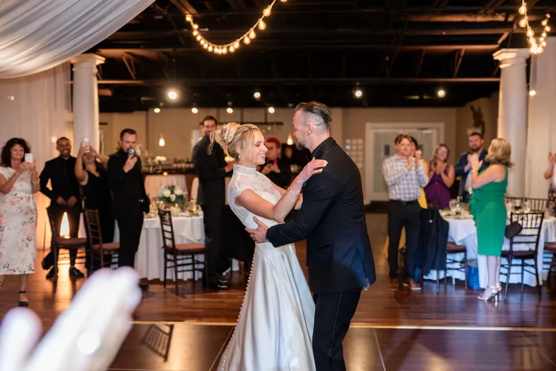 Two people dance together on a wooden floor beneath string lights while guests watch from the perimeter of an indoor venue.
