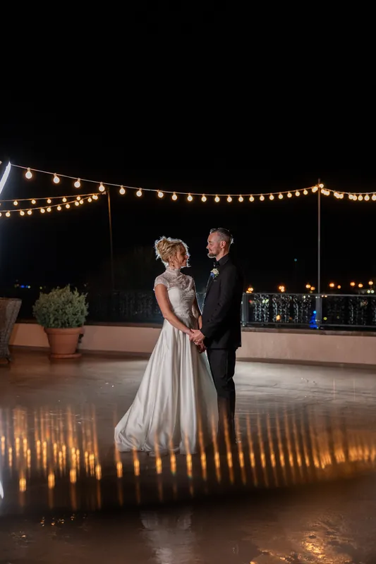 Under draped string lights on an outdoor terrace, two people in formal attire hold each other close during an evening first dance.