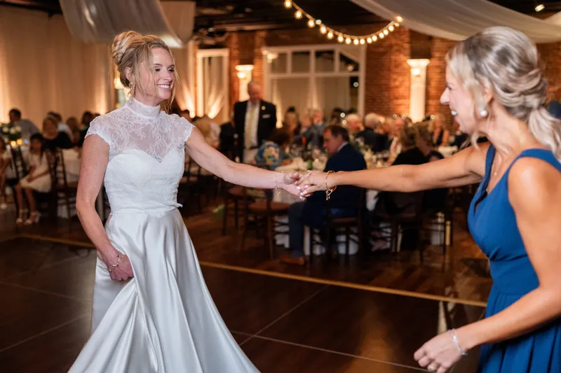 Someone in a flowing light gown reaches toward a guest in a blue dress during a indoor reception celebration.