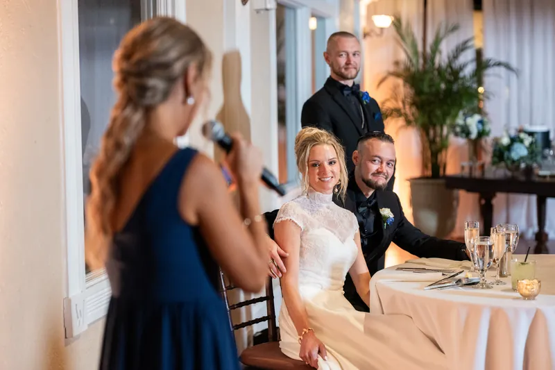 Wedding guests interact near a decorated table while two people in formal wear stand nearby in a warmly lit venue.