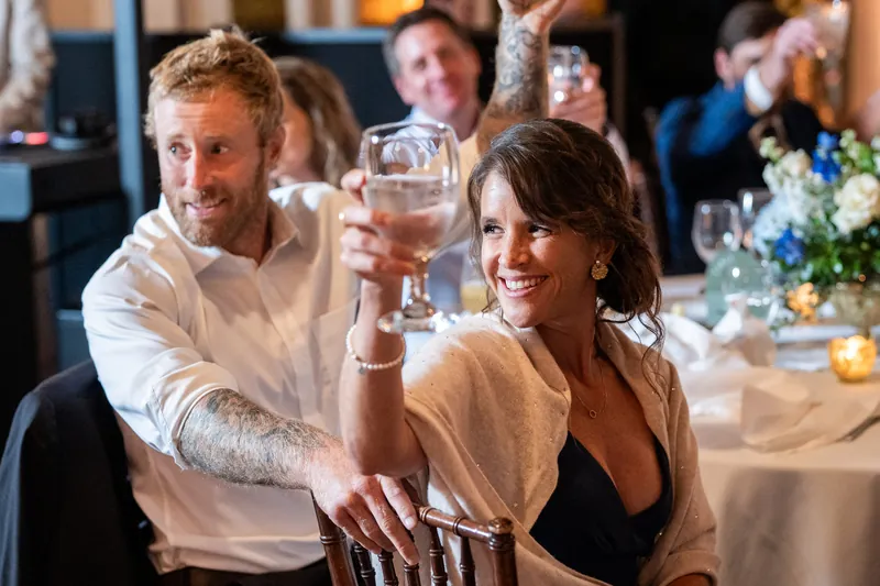 Guests raise glasses during a reception toast, smiling and toasting at a long table set with white linens.