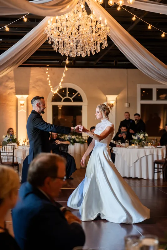 Two people in formal attire exchange vows on a dance floor beneath a crystal chandelier and draped ceiling while seated guests watch.