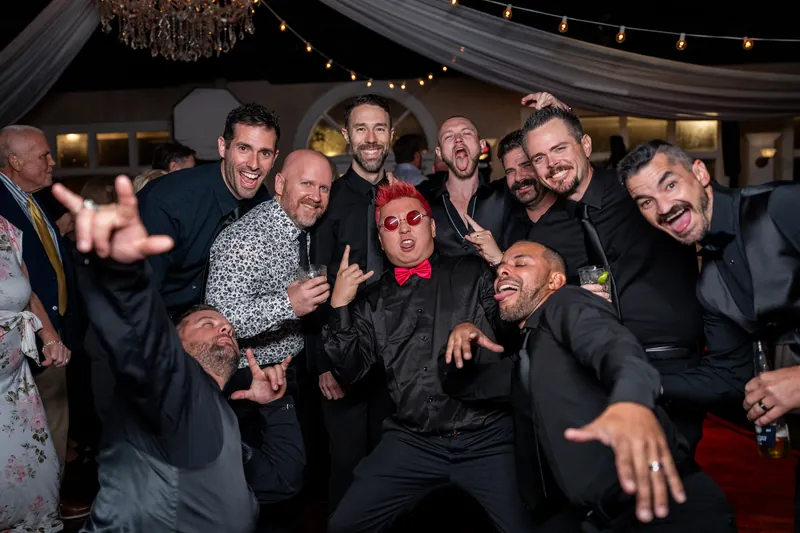 A group of men in dark suits pose together, smiling and gesturing playfully at the camera during the celebration.
