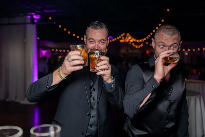 Two men in suits toast with drinks, their expressions animated as they clink glasses in front of purple uplighting.