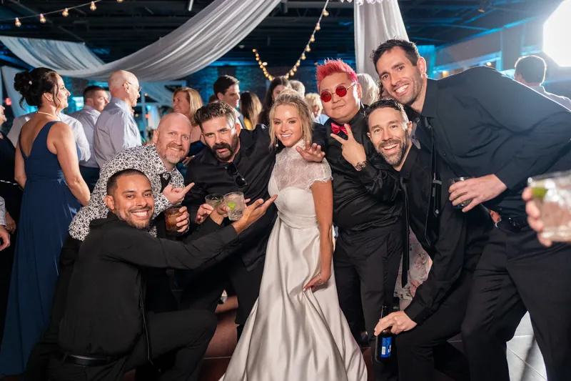 Li and Cody stand together in the center of a group of guests who embrace them both, all smiling for the camera under string lights.