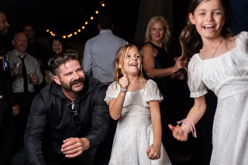 A man in a dark shirt dances with a young child in a white dress while guests watch under warm string lights.