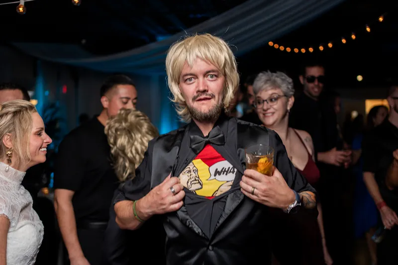 A man with blonde hair wearing a black jacket holds up colorful gift boxes during an indoor celebration with string lights overhead.