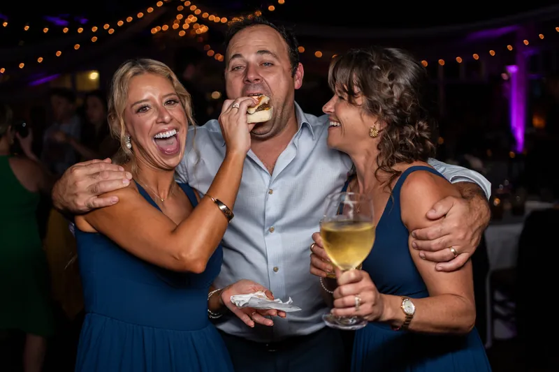 Two people embrace and laugh while holding drinks at a lively evening reception with string lights and purple ambient lighting.