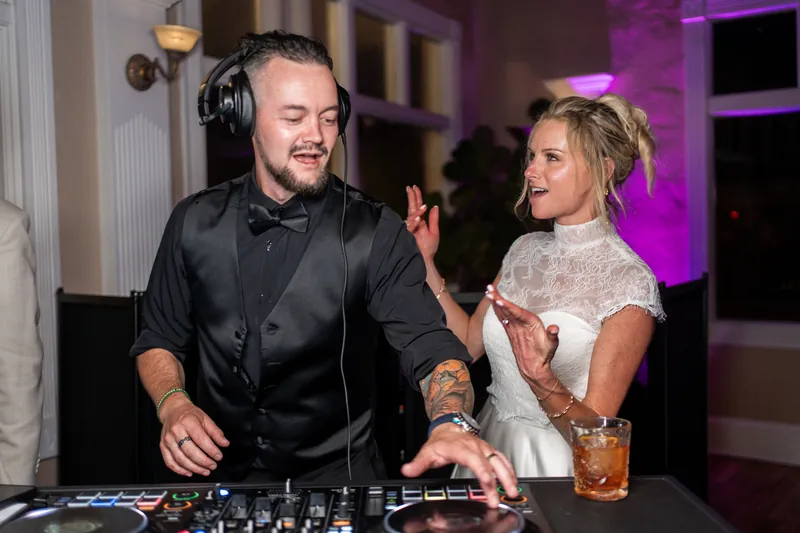 A DJ in headphones and black shirt chats with a woman in a white lace dress holding a cocktail at the dance floor.