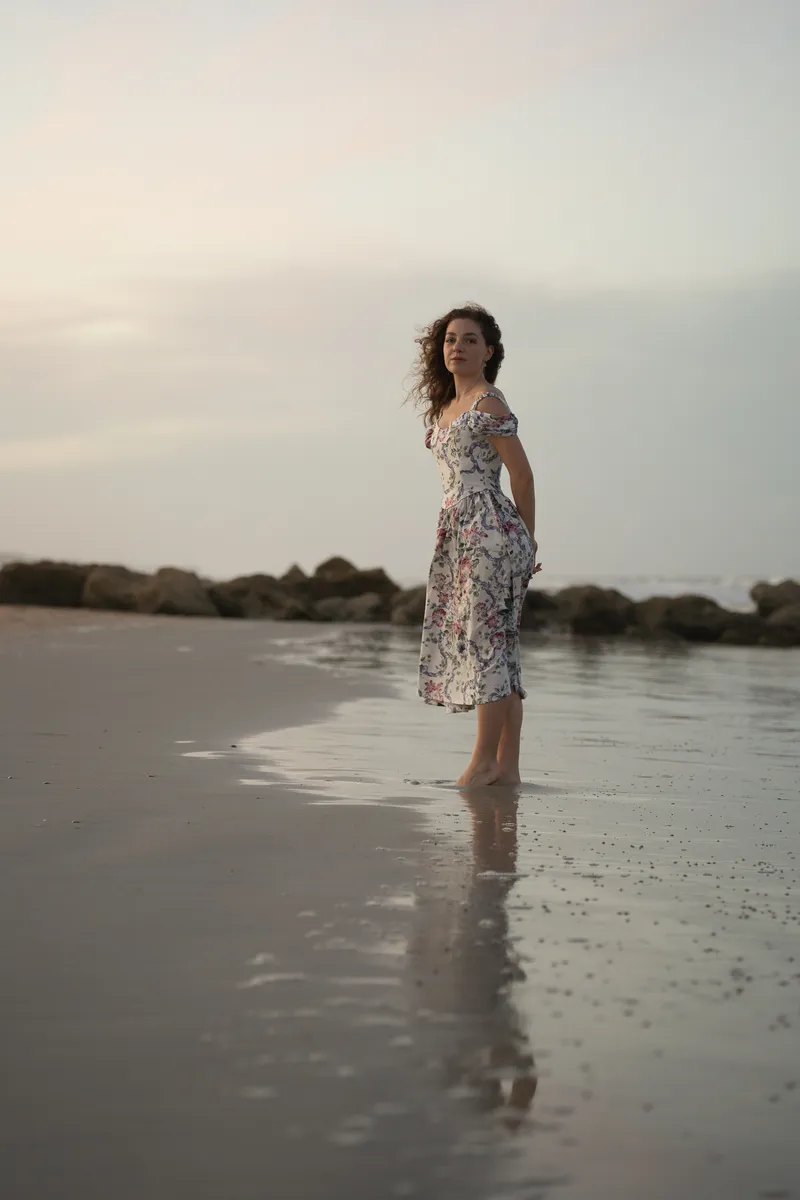 Lydia stands ankle-deep in shallow water at sunset, looking back toward the camera in her patterned dress.
