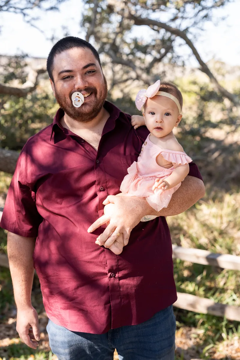 Dad holding baby girl with a pacifier in his mouth, laughing under the oak trees