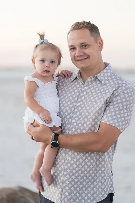 A man in a patterned shirt holds up a small child in a white dress, both smiling at the camera near the water.