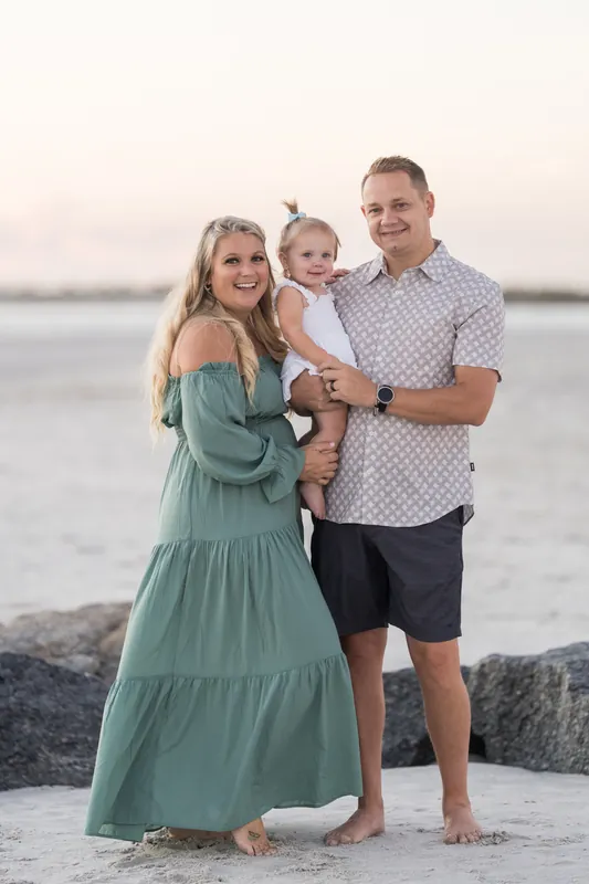 Two adults and a toddler pose barefoot on rocks at Vilano Beach as light fades, the ocean pale behind them.