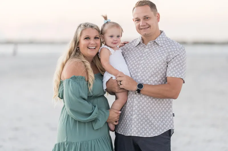 Two adults and a small child pose together on the beach at Vilano Beach, St. Augustine, smiling toward the camera as the sun sets over the water.