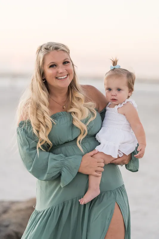 A person in a tiered sage dress holds a toddler in white, smiling at the camera with the beach and water beyond.