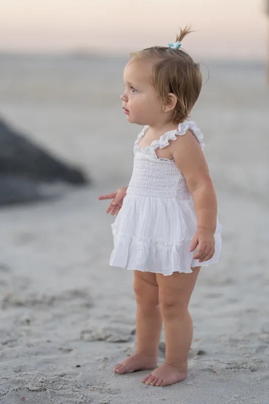 Toddler in a white sleeveless dress stands alone on the sand at Vilano Beach, St. Augustine, gazing toward the water at sunset.