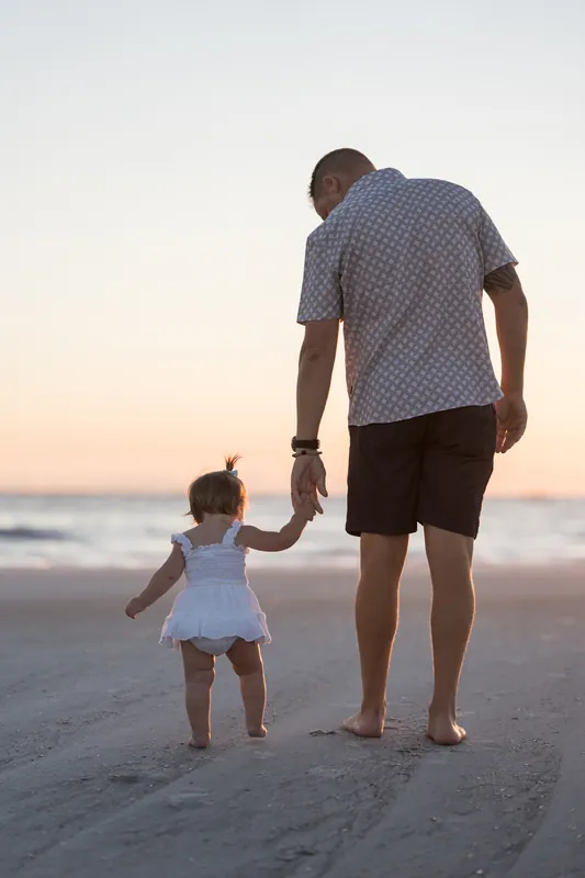 A man and small child hold hands on the sand at sunset, the child in a white dress and the man in a blue patterned shirt.