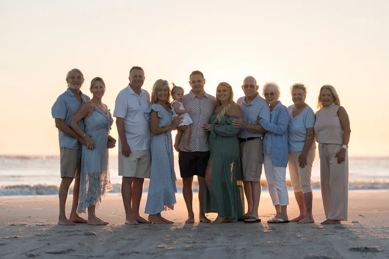 Extended family group of eleven people stands together on the beach at golden hour, dressed in shades of blue and green.