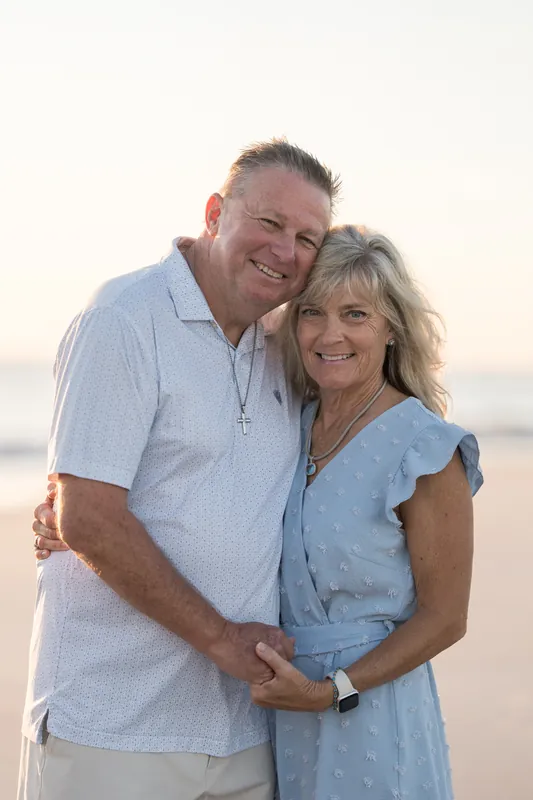 Mac and Danny embrace on the sand with the ocean behind them at sunset, both in light blue button-ups.