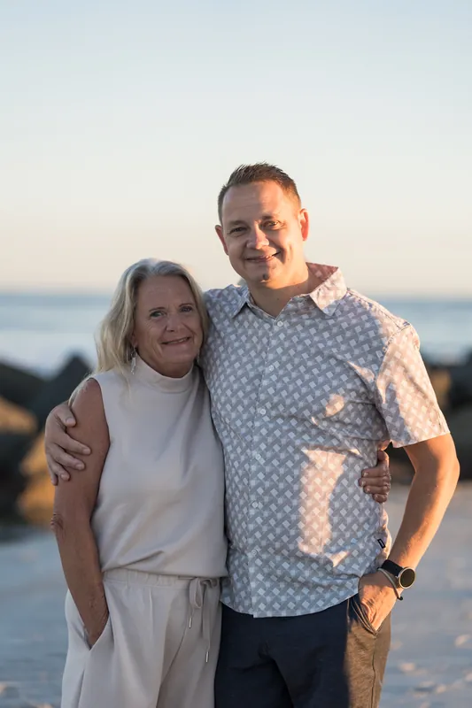 Mac and Danny stand arm-in-arm on the beach, smiling at the camera with soft evening light at their backs.