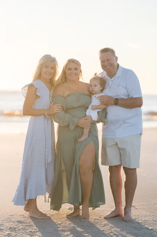 Four family members pose together on the sand—two women, a man in a light blue shirt, and a toddler in the center.