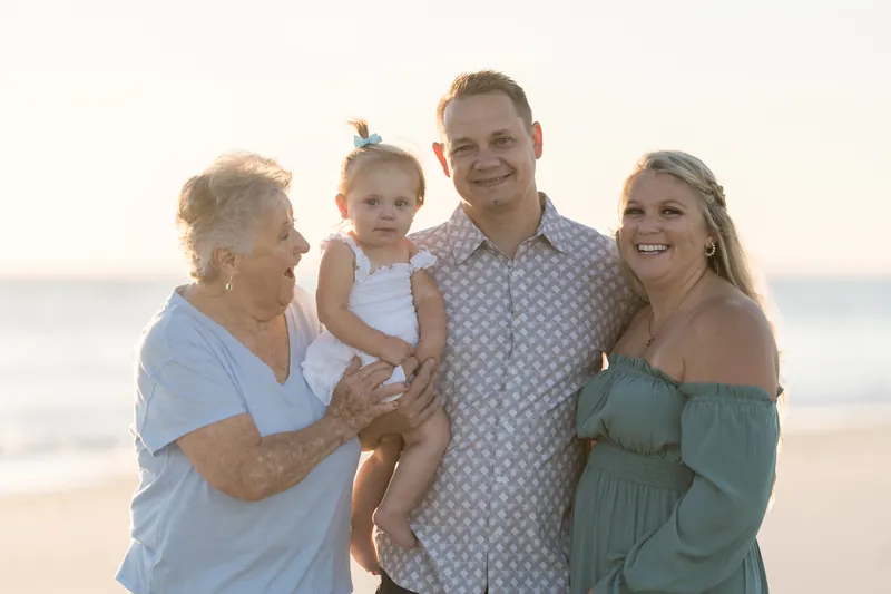 A group of five smiles at Vilano Beach, with one person holding a small child in the center and ocean surf visible behind them.