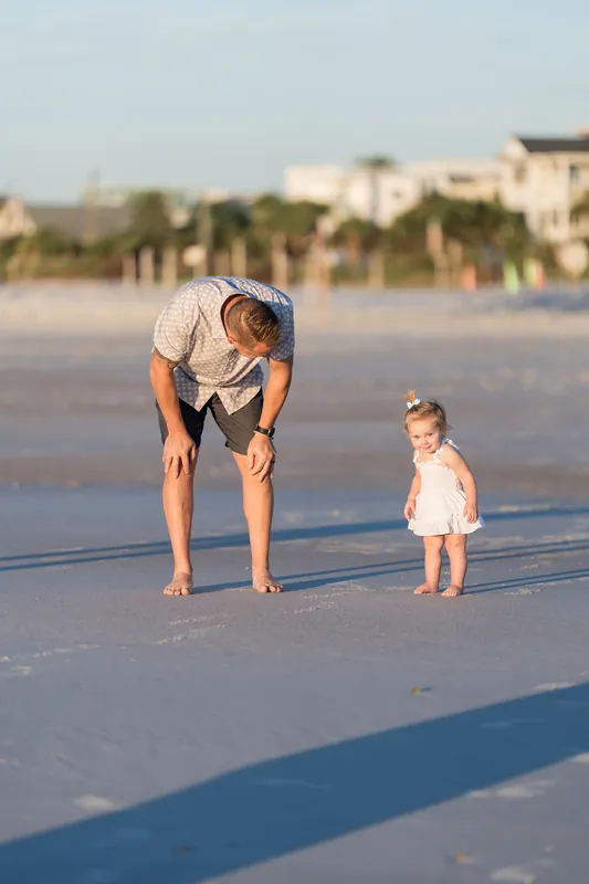 An adult bends down to play with a toddler in a white dress at the water's edge on Vilano Beach, with beach homes visible along the shore.