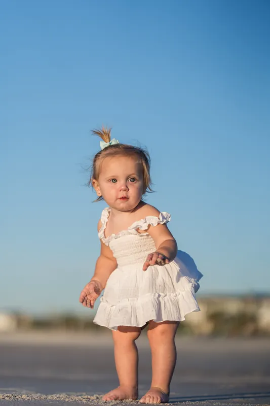 A toddler in a white ruffle dress stands alone on the wet sand at Vilano Beach, looking back toward the camera with the ocean behind.