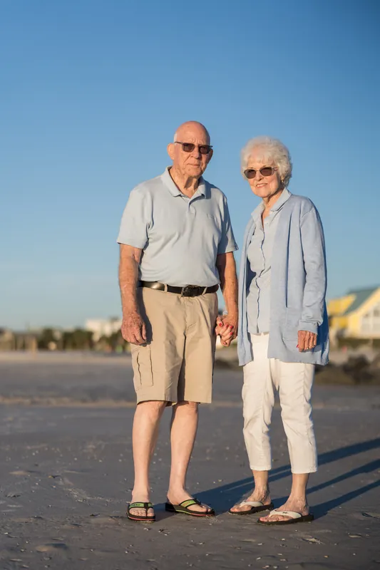 Two adults wearing sunglasses walk hand in hand across the beach at Vilano Beach, with calm surf and beach architecture visible in the background.