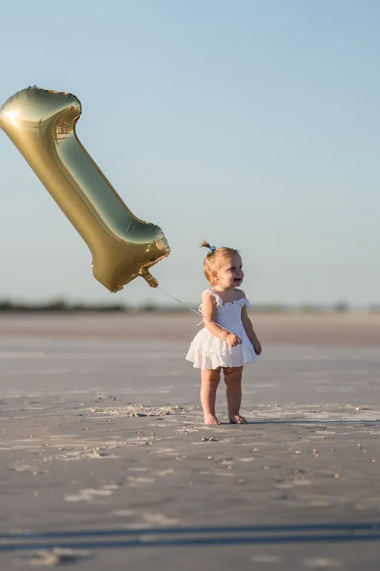 A toddler in a blue and white dress looks up at a tilted gold number 1 balloon on the beach.