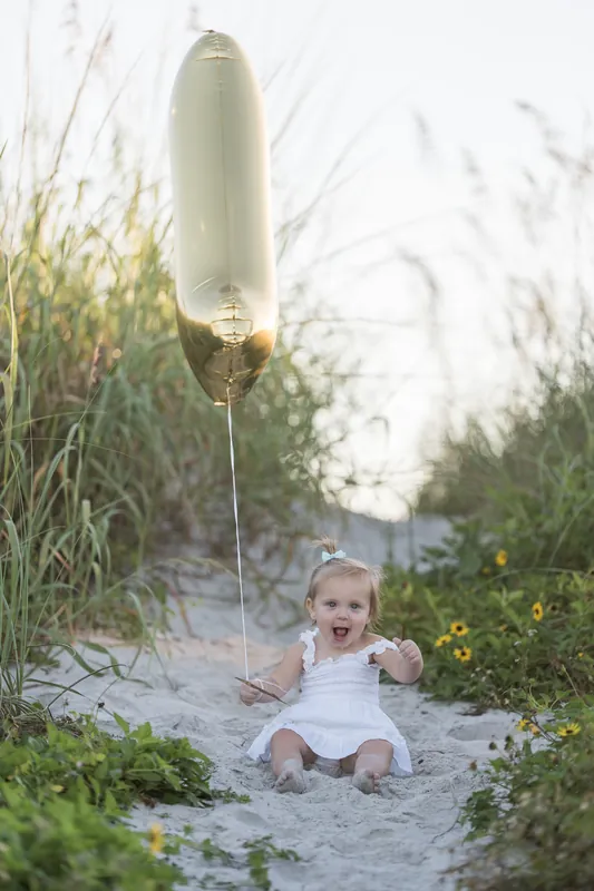 A smiling baby in a white dress holds the string of a gold number 1 balloon planted in the sand among dune grasses.