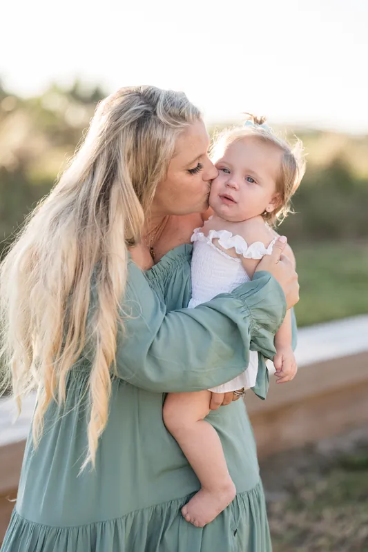 A woman in a sage green dress kisses a baby on the cheek, both smiling in golden afternoon light.