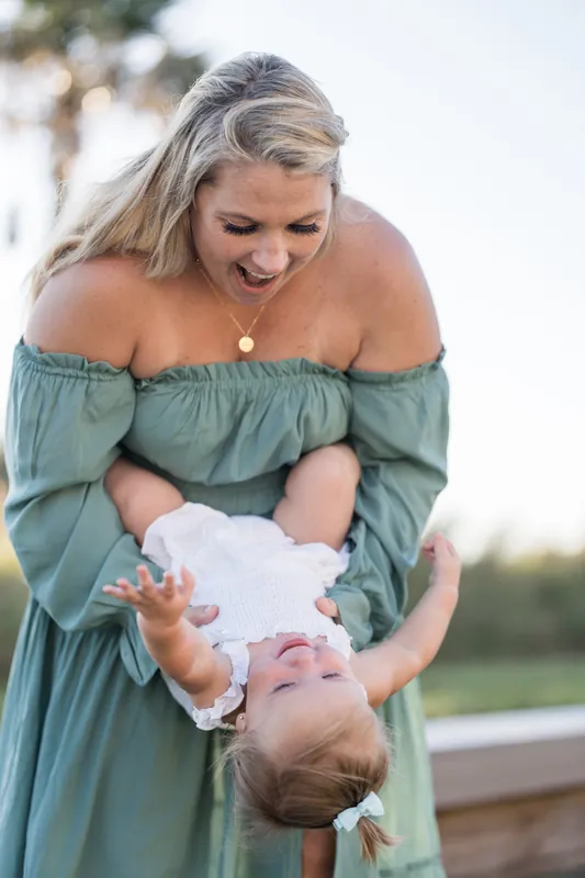 Person in sage off-shoulder dress holding a laughing toddler at Vilano Beach, with palms and water behind them.