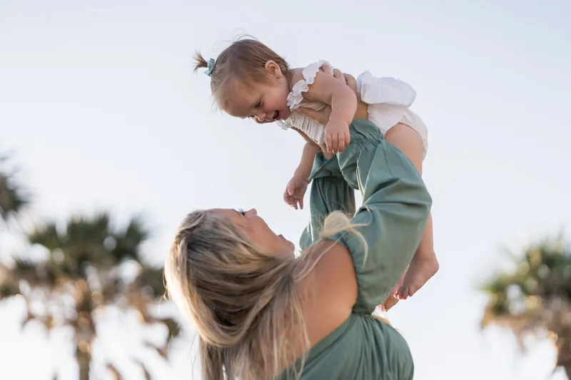 Adult lifting a small child overhead while smiling at Vilano Beach, with palm trees framing the scene.
