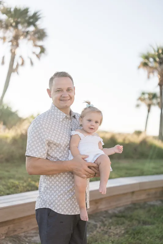 Adult in a patterned shirt cradling a toddler with arms outstretched at Vilano Beach, palms visible in soft golden light.