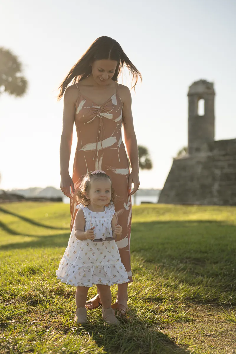 A woman in a tan bikini top and white patterned skirt holds a young child in a white dress while standing in a grassy field with historic ruins in the background.